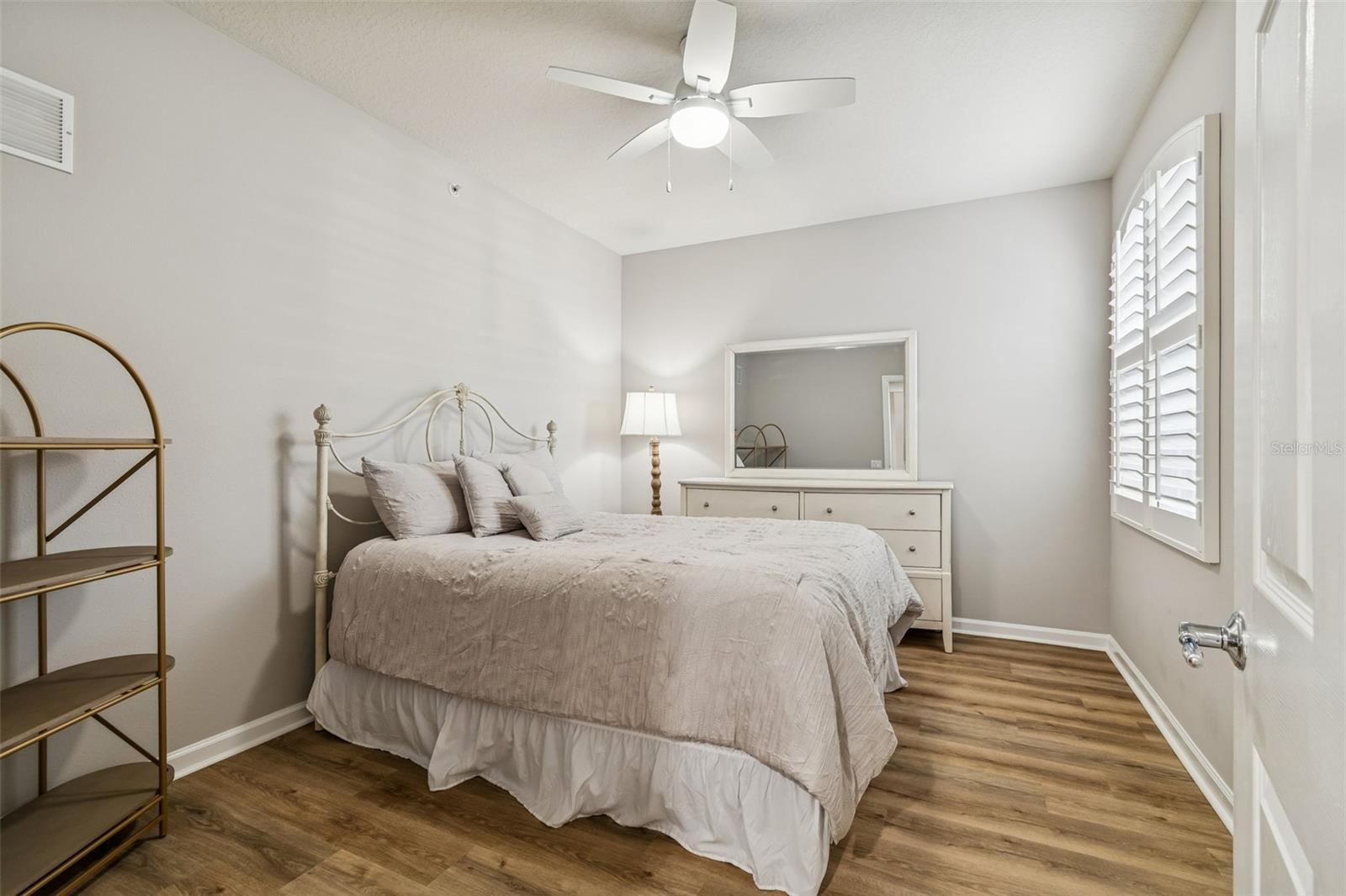 This cozy guest bedroom features plantation shutters, a ceiling fan, and warm-toned flooring, ideal for relaxation or hosting visitors.
