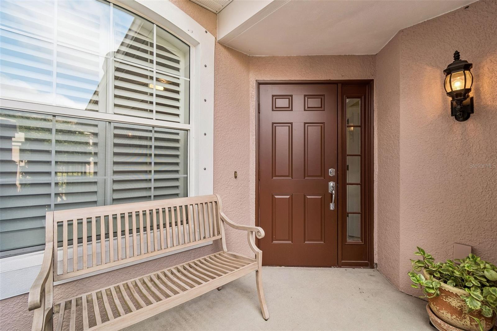 This inviting covered entry features a wood bench, wall-mounted lantern lighting, and a rich-toned front door with side glass paneling.