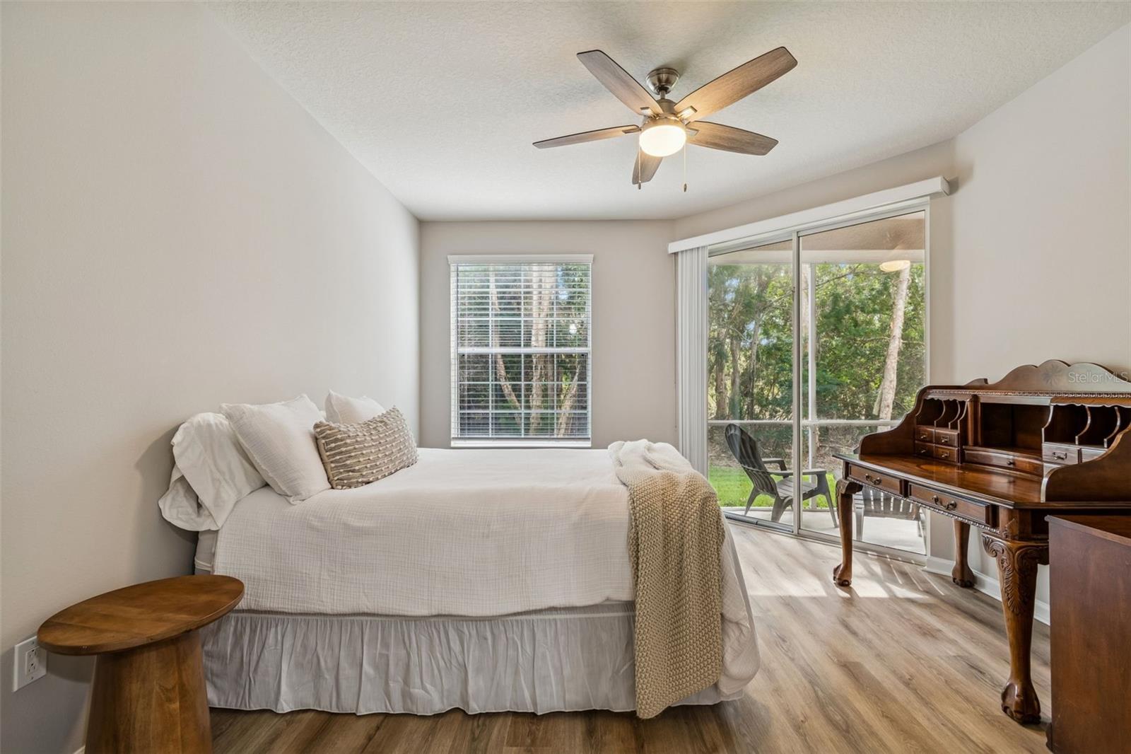 The serene primary bedroom welcomes you with soft natural light, neutral tones, and a peaceful view of the lush outdoors through expansive windows and sliding glass doors.