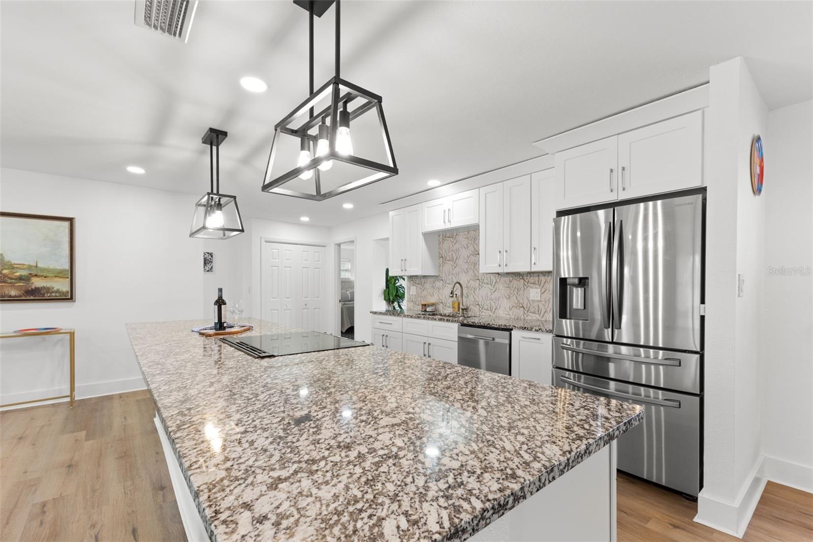 Expansive granite peninsula with overhang provides generous prep space and room for casual seating. White shaker-style cabinetry with brushed nickel hardware offers abundant storage, while contemporary pendant lighting adds architectural interest above. Recessed ceiling lights ensure bright, even illumination throughout the space. Continuous luxury vinyl plank (LVP) flooring enhances durability and flow into adjoining living areas.