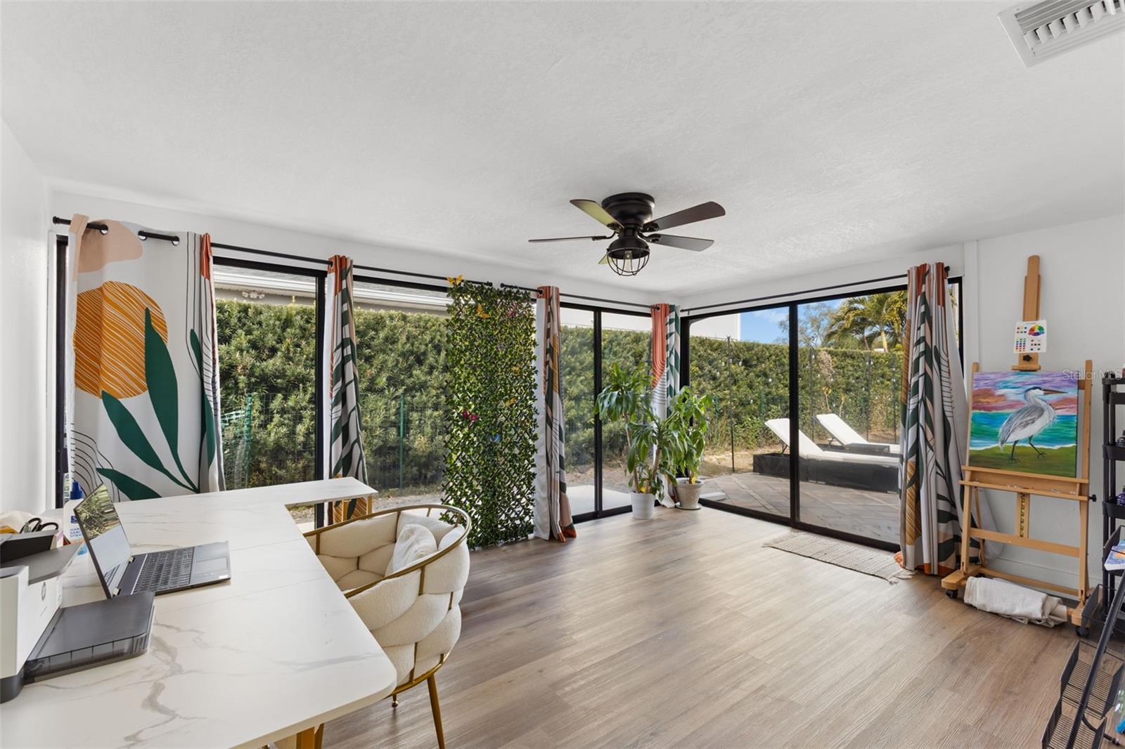 Bright and airy sunroom featuring expansive sliding glass doors that open directly to the rear paver patio, creating seamless indoor-outdoor living. Wood-look LVP flooring continues through the space, complemented by a modern ceiling fan. Large windows flood the room with natural light while offering private hedge-lined views.