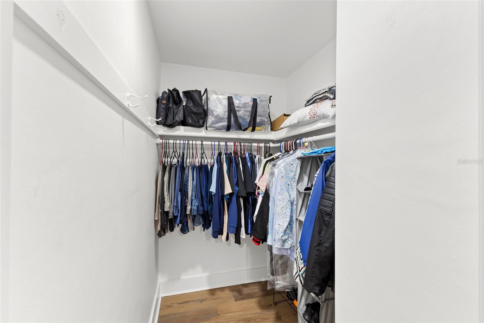 Walk-in primary closet offering generous hanging space with upper shelving for additional storage. Functional layout provides room for clothing organization while maintaining open floor space. Bright white walls and wood-look flooring continue the home’s clean, cohesive finish. Ideal for everyday convenience and efficient wardrobe storage.