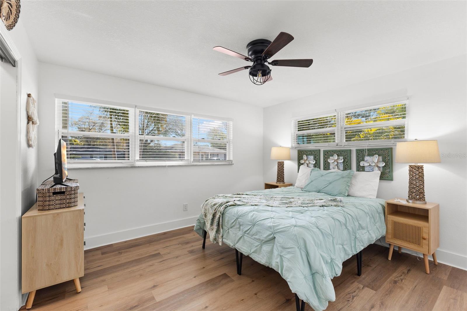 Light-filled secondary bedroom featuring wide-plank LVP flooring, crisp white walls, and multiple windows with plantation-style blinds that invite in natural light while maintaining privacy. A modern ceiling fan adds comfort year-round.