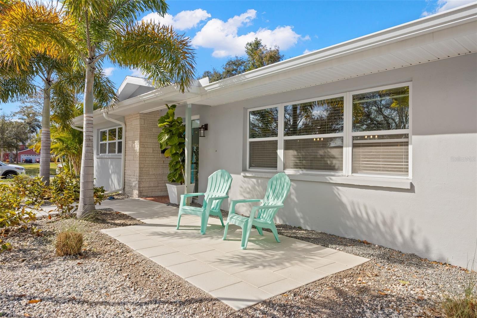 Spacious covered front patio with extended paver-style tile surface, offering a defined outdoor seating area beneath a wide roof overhang with finished soffits and gutter system. Large front-facing windows with white trim enhance curb appeal while providing generous natural light to the interior. The landscaped front yard features decorative gravel and mature tropical plantings for low-maintenance appeal, complemented by a defined walkway leading to the main entrance.