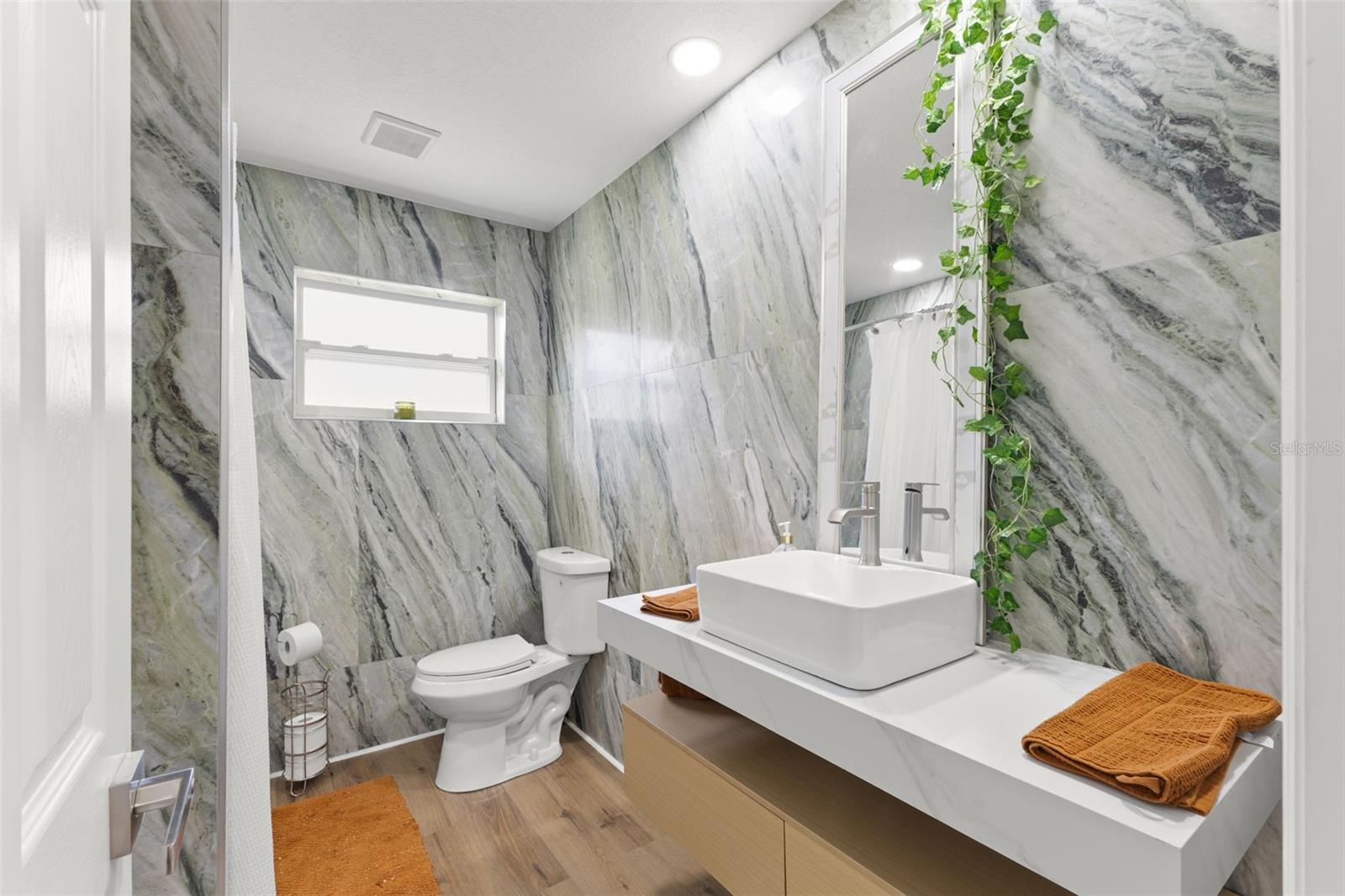 Beautifully updated second bathroom featuring dramatic large-format tile walls with elegant natural veining, creating a spa-inspired feel. The laminate countertop is paired with a modern vessel sink and sleek brushed-nickel faucet, offering clean lines and contemporary style. LVP flooring adds warmth and durability, while the oversized mirror enhances light and space.