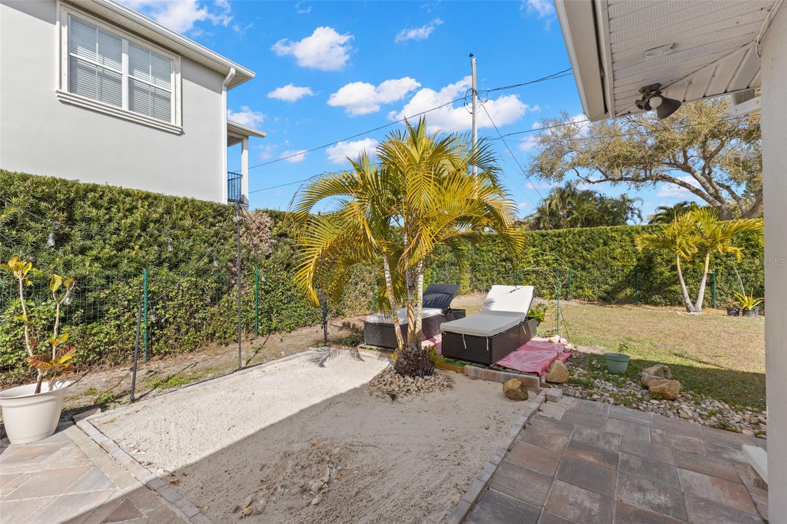 Generous outdoor living space featuring defined patio zones and open grassy area. Mature hedge line enhances privacy and buffers neighboring properties. Tropical plantings and palm accents reinforce the inviting backyard atmosphere, while the paver surface provides a durable, low-maintenance foundation ideal for everyday enjoyment and entertaining.