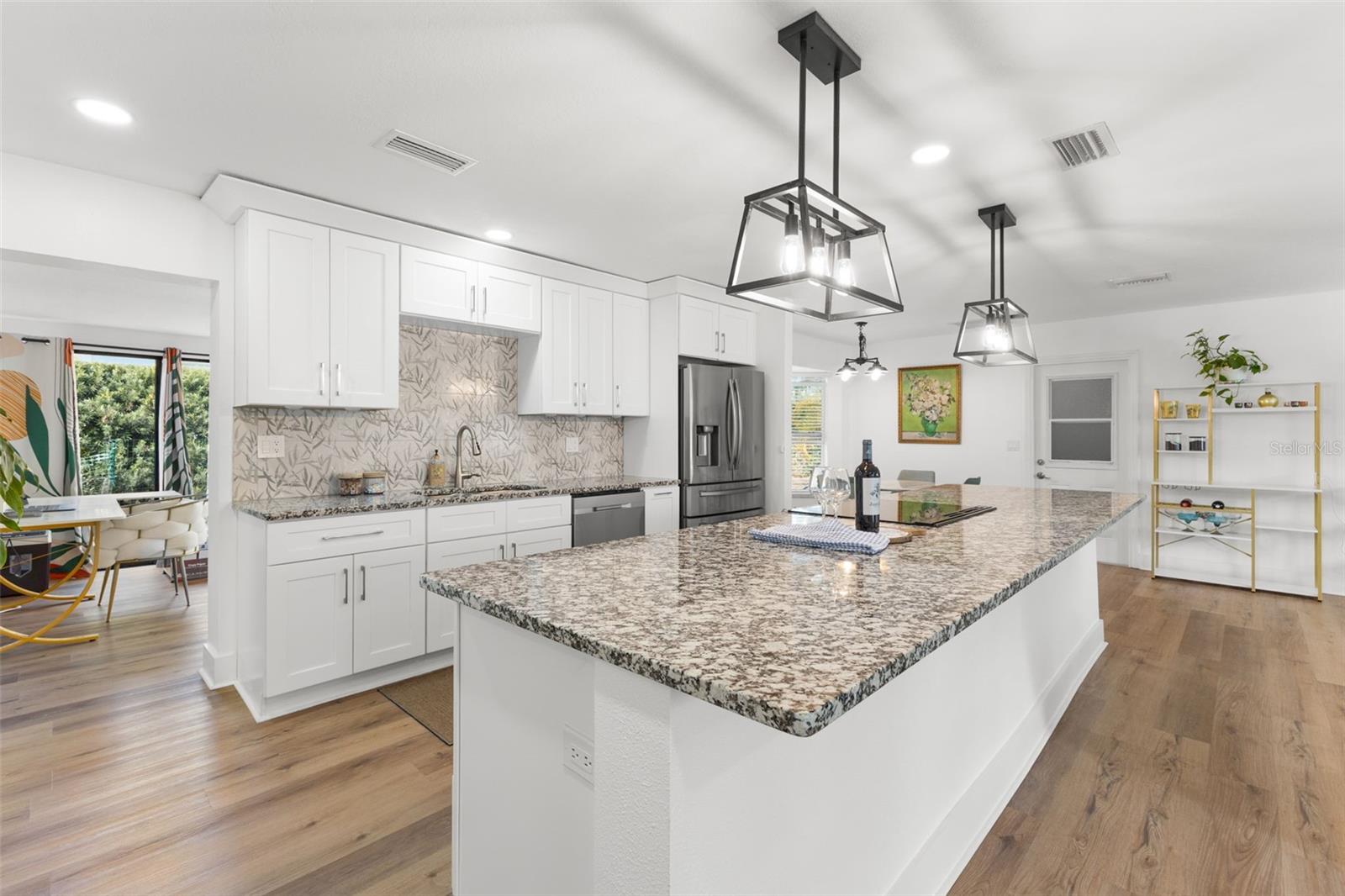 Striking oversized granite island anchors the kitchen, offering expansive prep space and additional storage below. Contemporary pendant lighting provides focused illumination, complemented by recessed ceiling lights for bright, even coverage throughout.White shaker-style cabinetry extends to the ceiling, maximizing vertical storage, and is paired with brushed nickel hardware for a clean, modern finish. Granite countertops continue along the perimeter, highlighted by a full-height decorative tile backsplash that adds texture and visual interest. Stainless steel French-door refrigerator, dishwasher, and undermount sink with gooseneck faucet complete the thoughtfully designed, highly functional workspace. Continuous luxury vinyl plank flooring enhances durability and seamless flow into adjoining living areas.