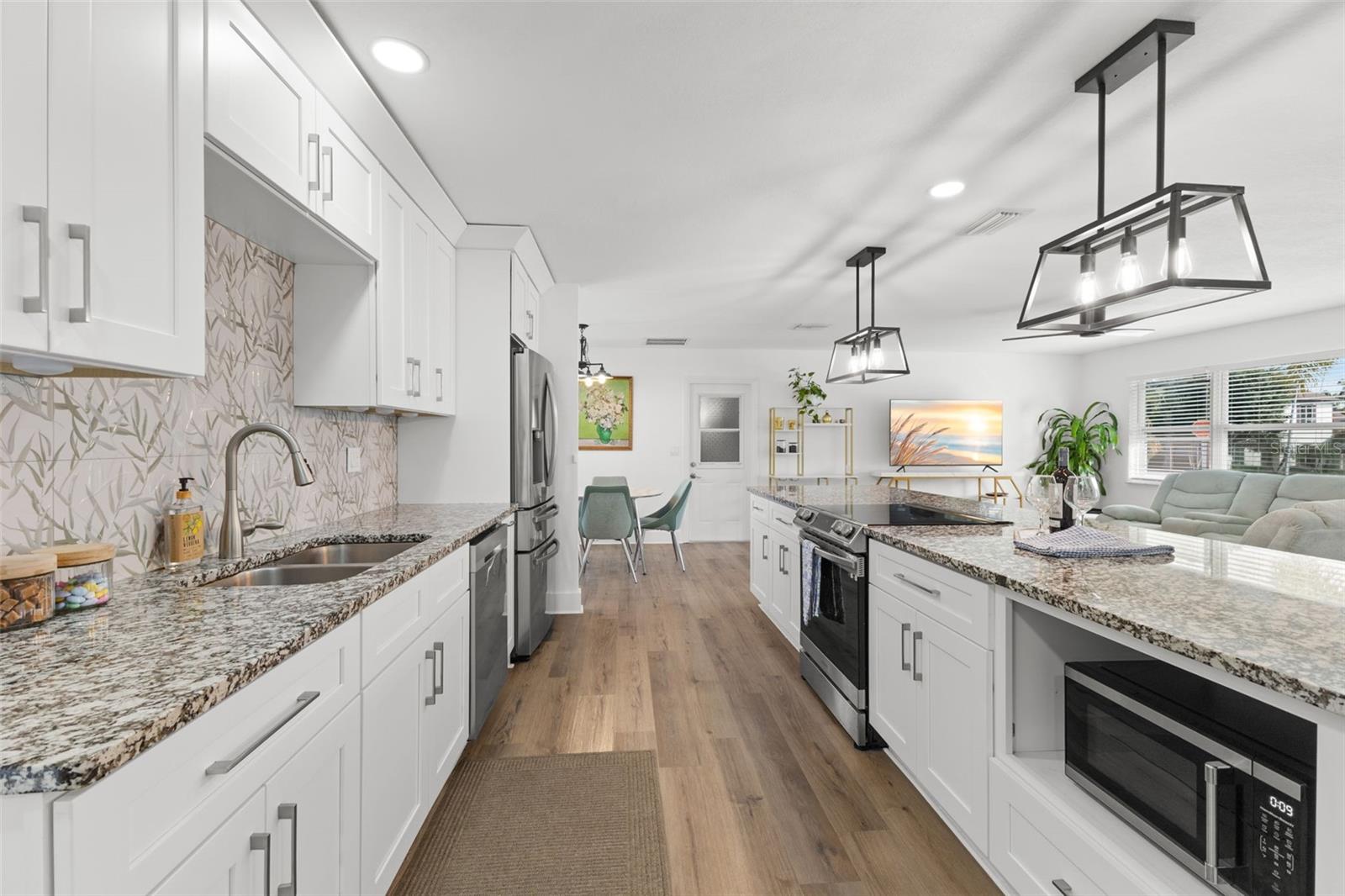 Functional galley-style layout with dual-sided cabinetry maximizes storage and efficiency. White shaker cabinets extend to the ceiling, providing additional upper storage. Granite countertops and full-height decorative tile backsplash add texture and visual interest. Stainless steel Samsung appliances, including French-door refrigerator and dishwasher, complement the clean, modern aesthetic. Undermount stainless sink with gooseneck faucet enhances workspace functionality.