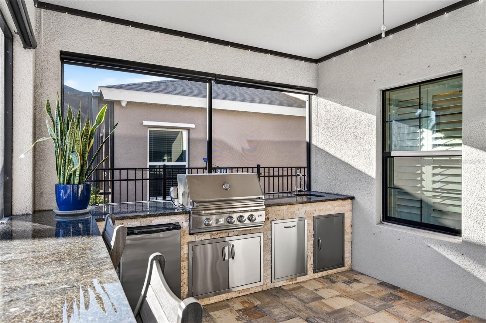 Outdoor kitchen under cover with granite countertops, grill, refrigerator, concealed trash bin and sink.