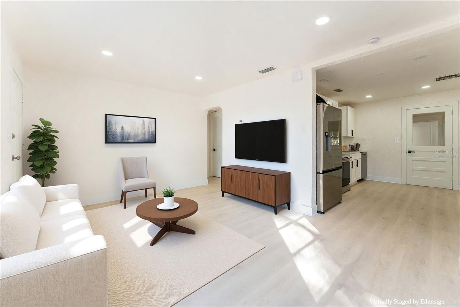 Spacious living area with direct sightlines into the kitchen, creating an open and connected feel. Recessed lighting, neutral tones, and wide-plank flooring give the space a bright, cohesive look. The open doorway to the kitchen enhances functionality and flow for gatherings. Image has been virtually staged to demonstrate potential furnishing and design.