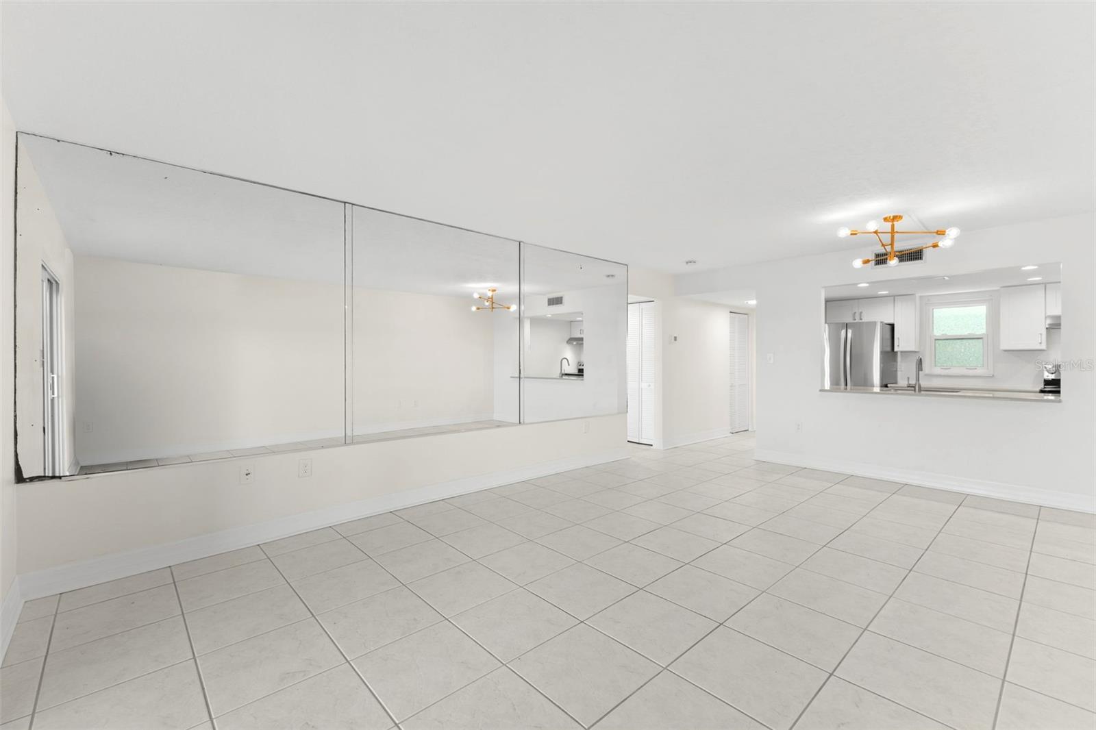 Bright, open-concept living and dining space enhanced by a wall of mirrors that expands the room visually, complemented by tile flooring and convenient kitchen access.