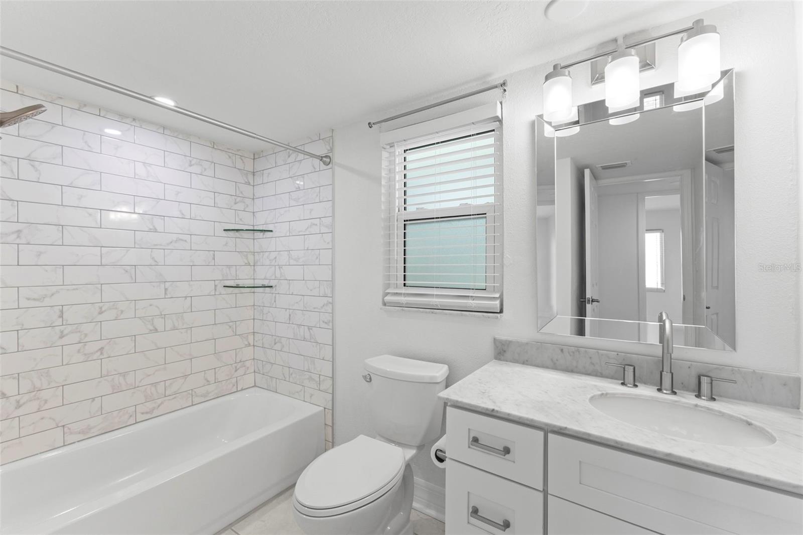 Updated bathroom featuring marble-look subway tile surround, glass shelving accents, and a sleek single-sink vanity with marble countertop.