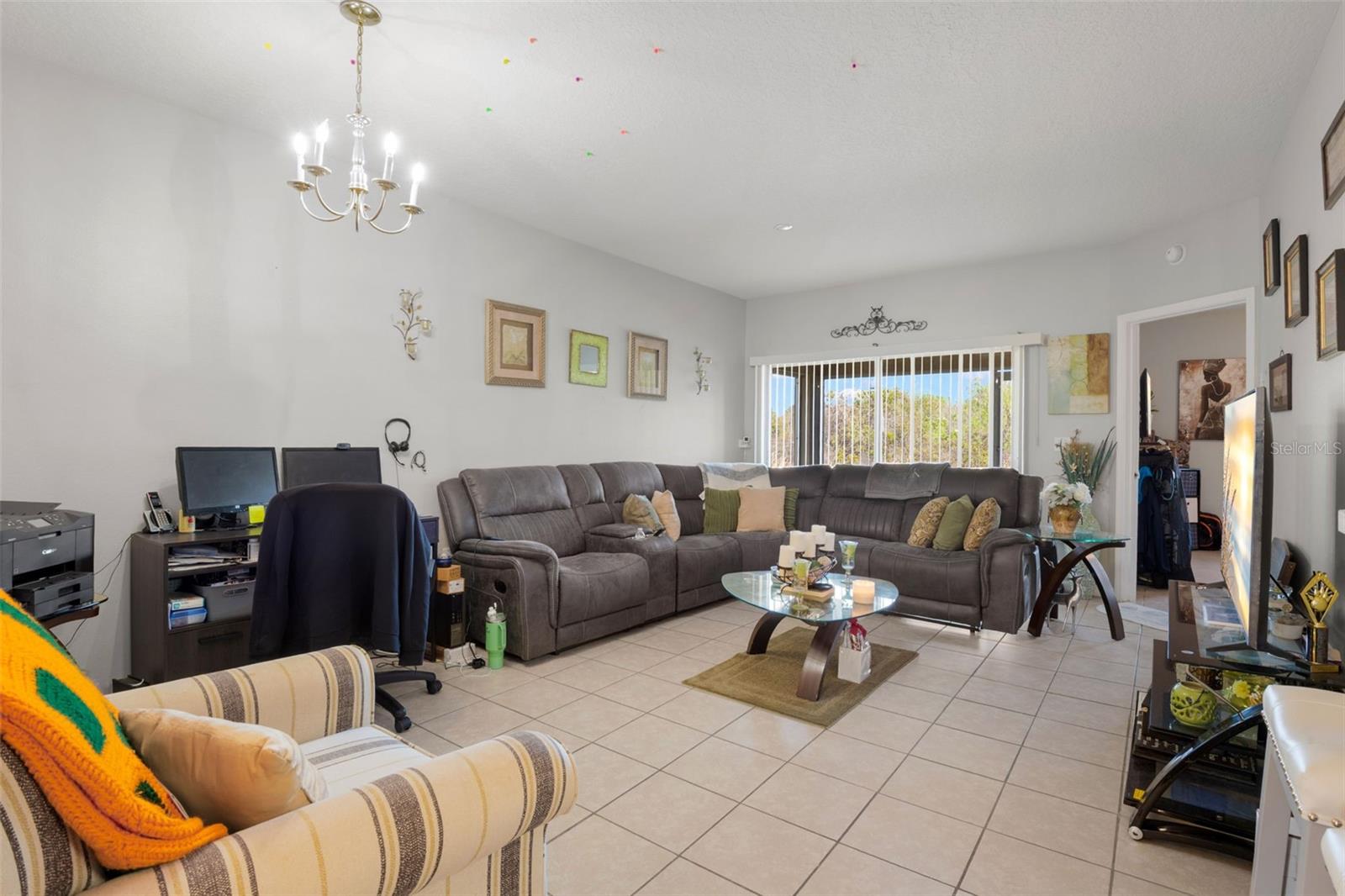 Spacious living area with ceramic tile flooring throughout and a bright, open feel. Sliding glass doors with vertical blinds allow natural light to fill the space while offering private views. The open-concept layout flows seamlessly into the dining area.