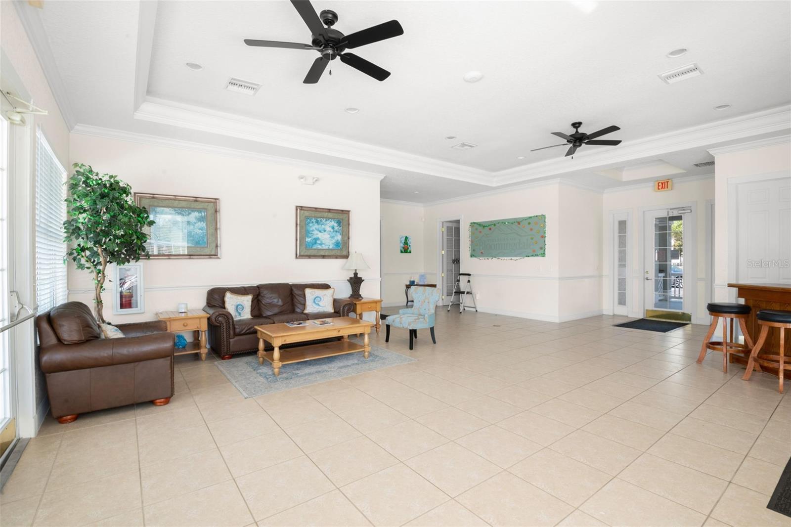 Spacious and inviting clubhouse interior featuring tile flooring, and ceiling fans throughout. Comfortable seating areas create a welcoming space for residents to relax or gather, while natural light fills the room through multiple windows and glass doors.