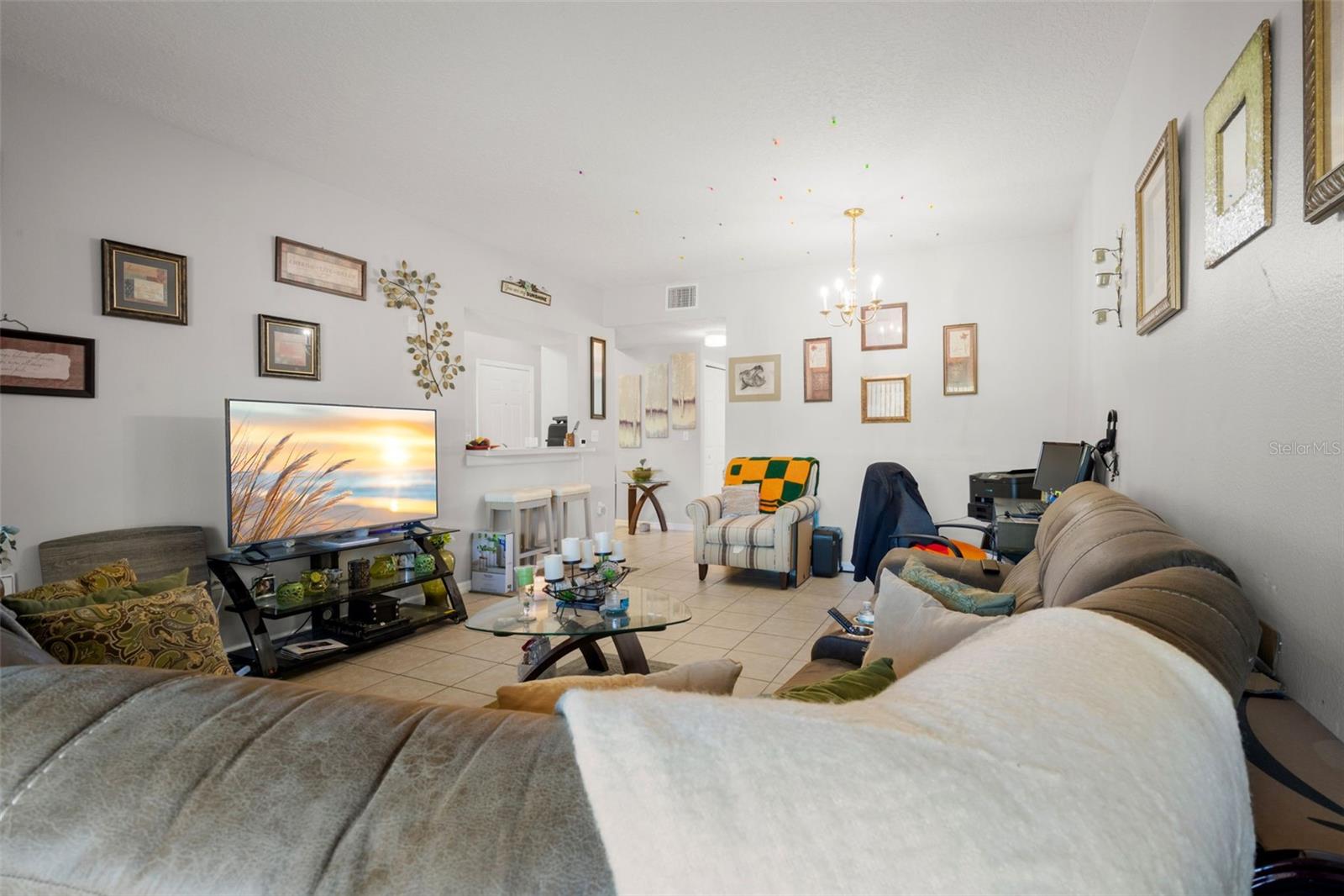 Open-concept living space with ceramic tile flooring throughout and a seamless flow into the dining area. The designated dining space is positioned beneath the chandelier, offering room for a dining table while maintaining connection to both the kitchen and living room. A breakfast bar with additional seating provides casual dining options.