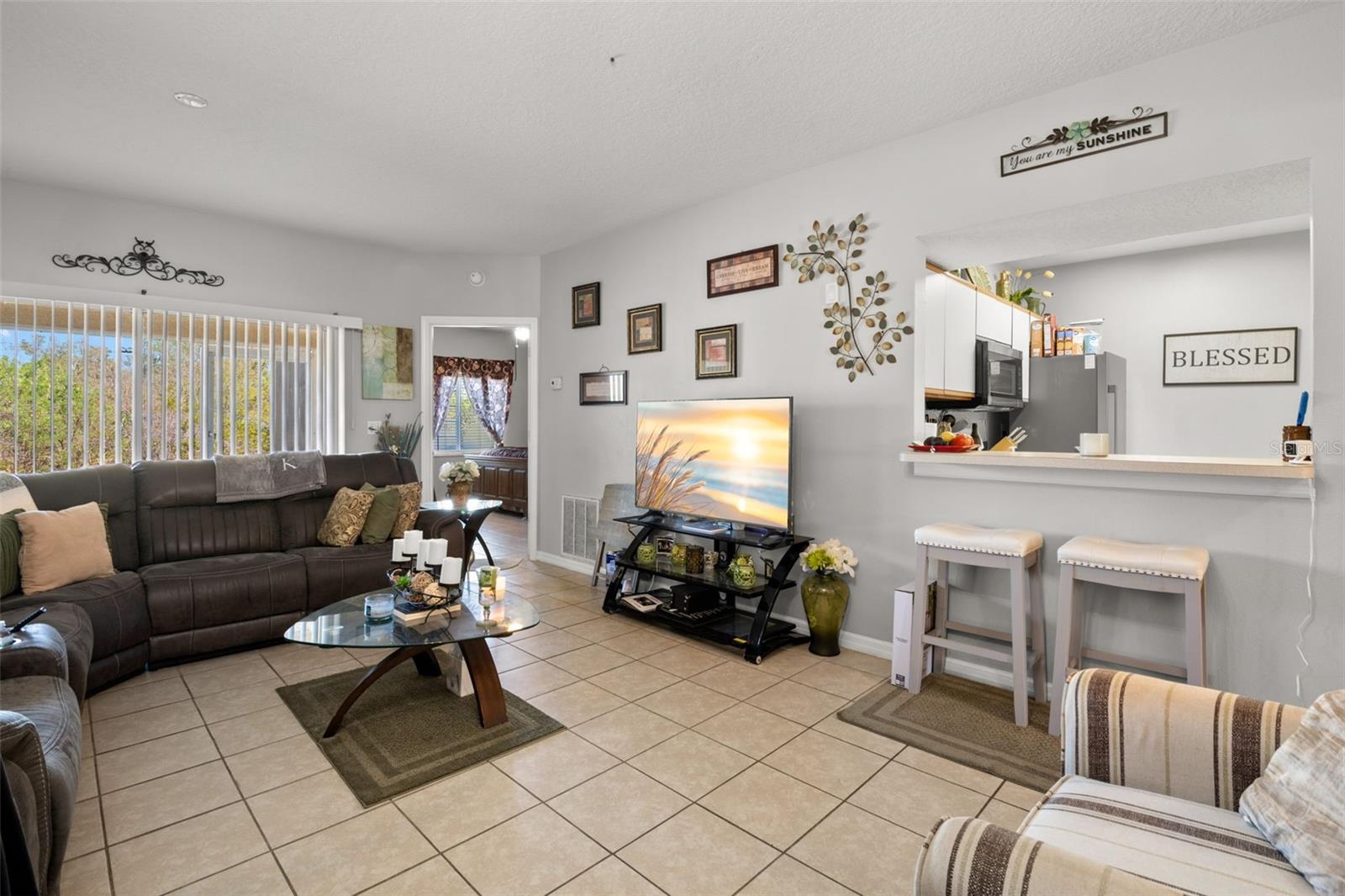 Bright, open main living area featuring ceramic tile flooring and a seamless connection to the kitchen. The breakfast bar offers additional seating and casual dining space while maintaining clear sight lines to the living room. Sliding glass doors provide natural light and access to the screened lanai. The split-bedroom floor plan extends off the main living area, offering privacy and functional flow throughout the home.