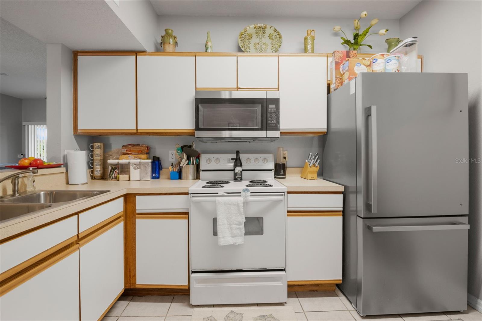 Functional kitchen layout with ample cabinetry and generous counter space for meal prep and storage. Features include range, microwave, refrigerator, dishwasher, and disposal, along with a double-basin sink and breakfast bar seating just beyond. Clean, neutral finishes and ceramic tile flooring offer a low-maintenance and versatile workspace that connects seamlessly to the main living area.