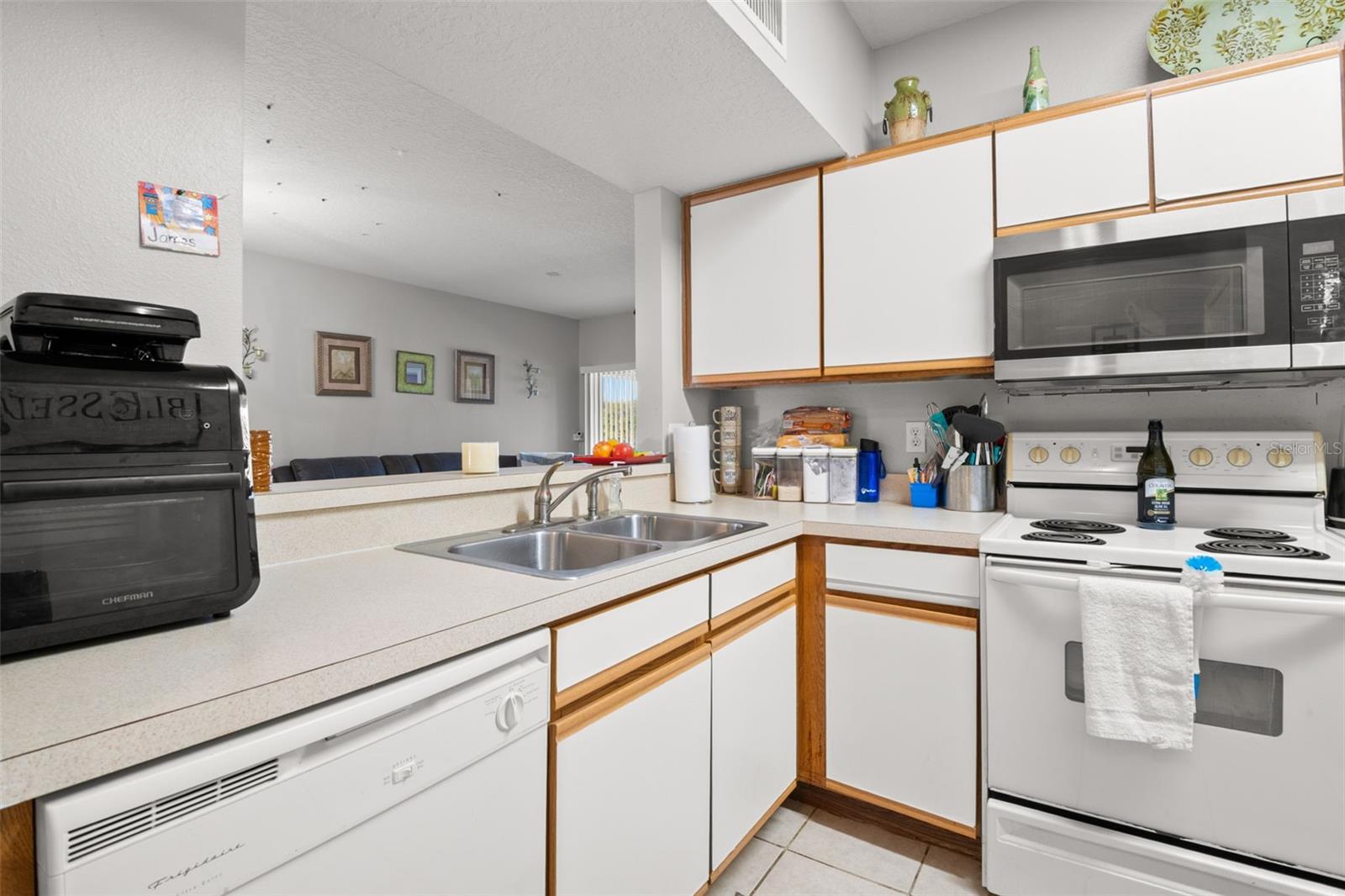 Functional L-shaped kitchen layout with double-basin sink, extended counter space, and clear sight lines into the living room. The breakfast bar provides additional seating and enhances the open-concept design, making the space ideal for both everyday meals and entertaining. Continuous ceramic tile flooring maintains a cohesive flow throughout the main living areas.