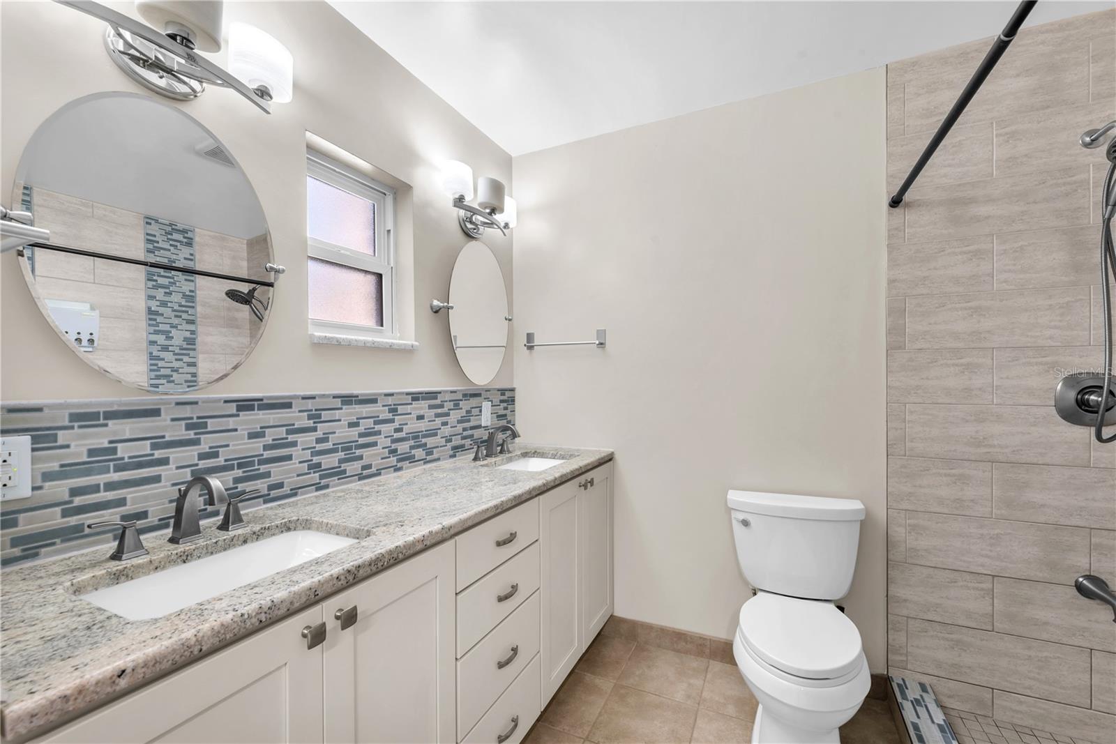 The spa-inspired en-suite bath features dual sinks set under solid stone counters, frameless adjustable mirrors, and white cabinetry with brushed nickle hardware.