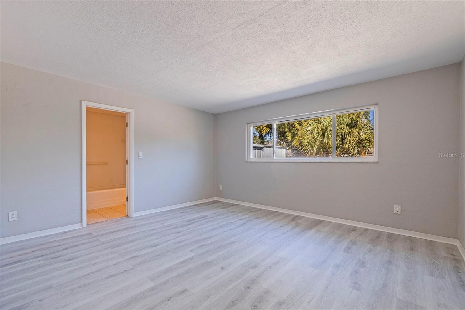 Primary bedroom featuring vinyl plank flooring and soft neutral tones for a calm, cohesive feel. A wide picture window brings in natural light and views of the backyard greenery, while the spacious layout easily accommodates a full bedroom suite. Direct access to the en suite bath adds everyday convenience and functionality.