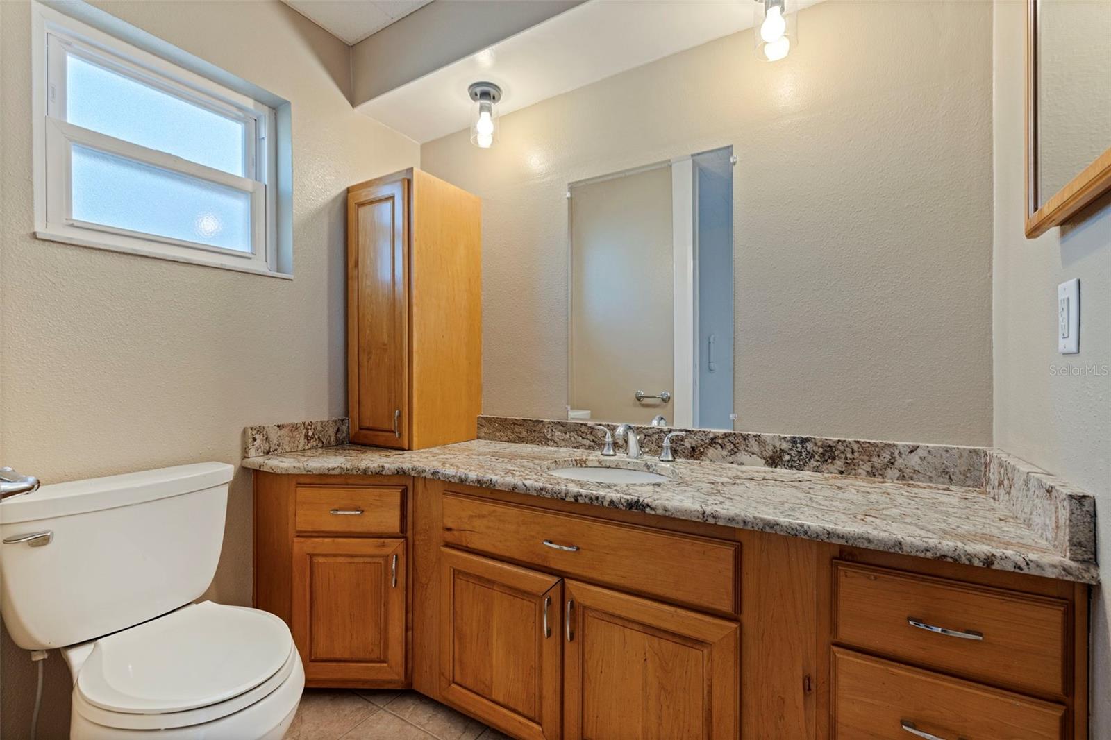 Primary bathroom featuring an extended granite countertop with integrated sink and abundant cabinet storage. Warm wood cabinetry provides generous drawer and cupboard space, complemented by updated hardware and neutral finishes. A frosted window allows natural light while maintaining privacy, creating a bright and functional en suite bath.