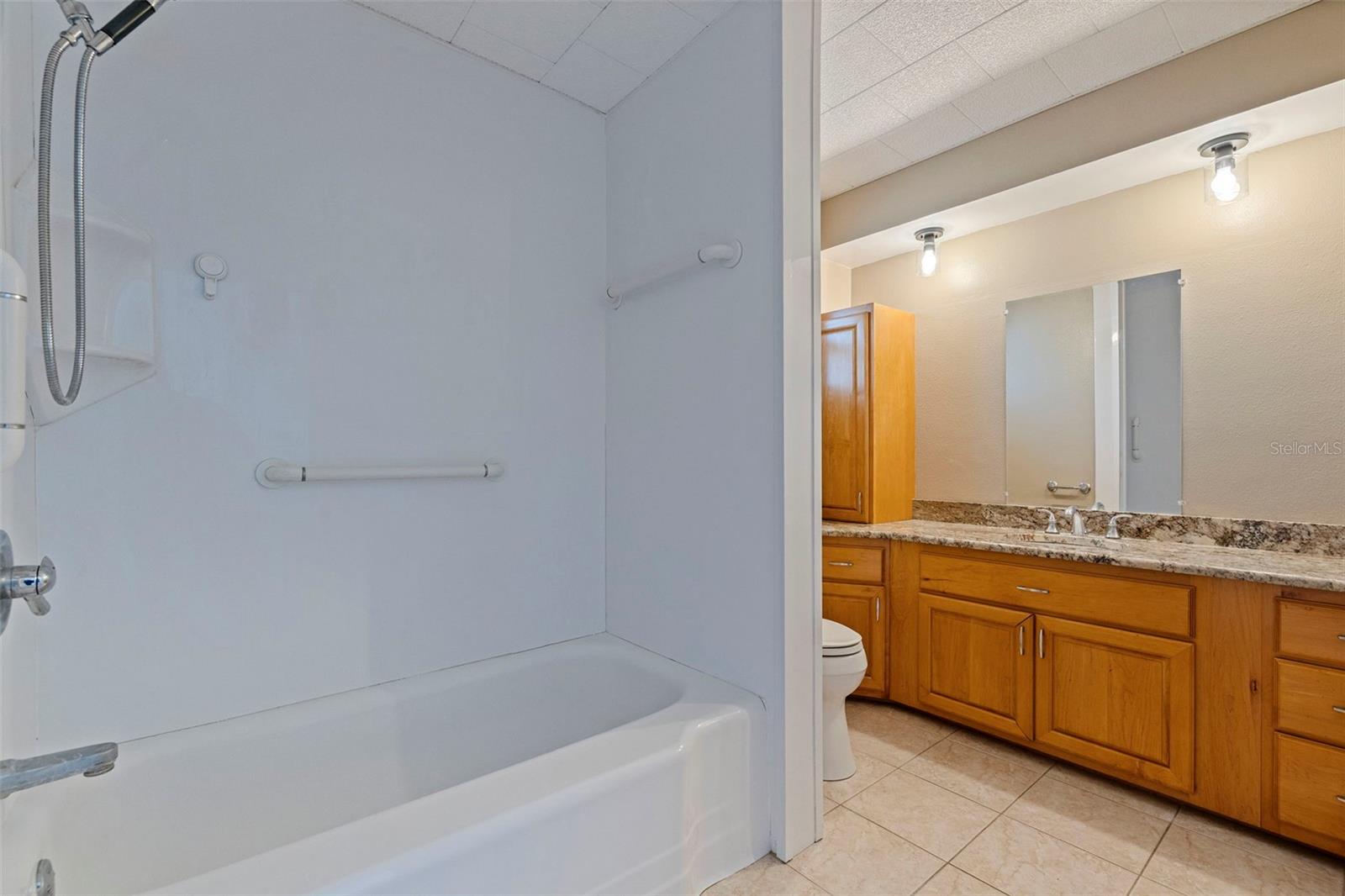 Primary bathroom with a full tub and shower combination featuring easy-care surround and built-in grab bars. The adjacent granite-topped vanity offers generous counter space and extensive wood cabinetry for storage. Neutral tile flooring and coordinated finishes create a clean, functional en suite layout.
