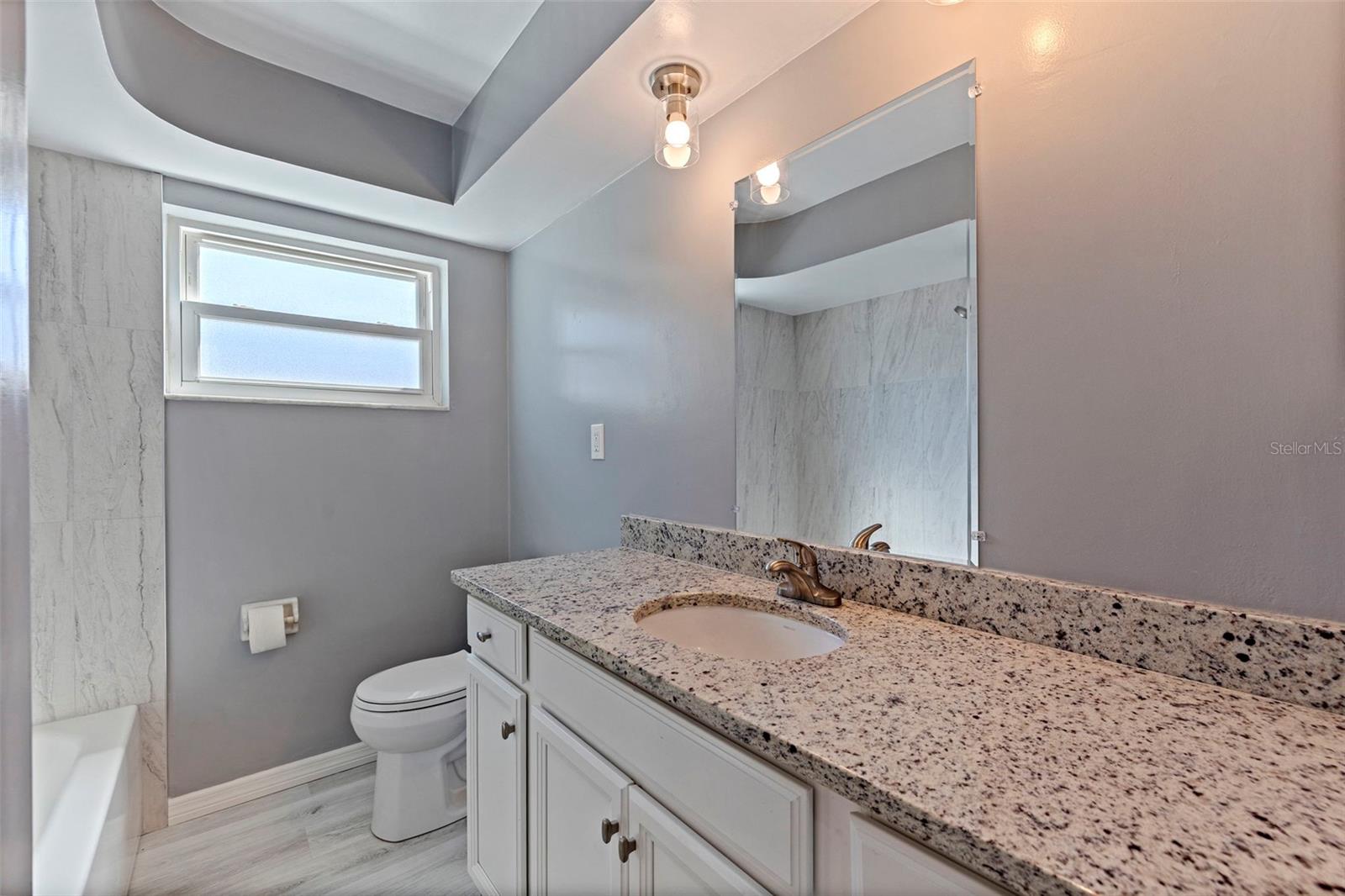 Second bathroom featuring a granite-topped vanity with undermount sink and updated fixtures. A tub and shower combination with marble-style tile surround adds a clean, modern touch, while the frosted window provides natural light with privacy. Neutral finishes create a bright and functional space for guests or family use.