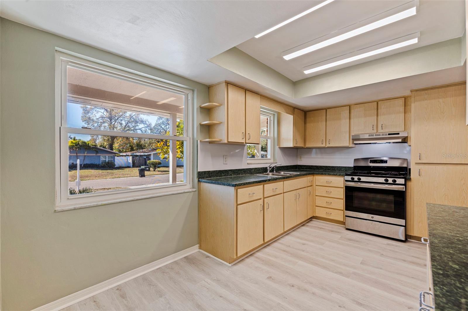 Light-filled eat in kitchen featuring abundant cabinetry, ample counter space, and a functional L-shaped layout. Large front-facing window provides natural light and neighborhood views. Equipped with a gas range, range hood, double sink, and pantry storage, the space offers practical workflow and everyday convenience. Updated vinyl plank flooring continues seamlessly from the main living areas for a cohesive interior finish.