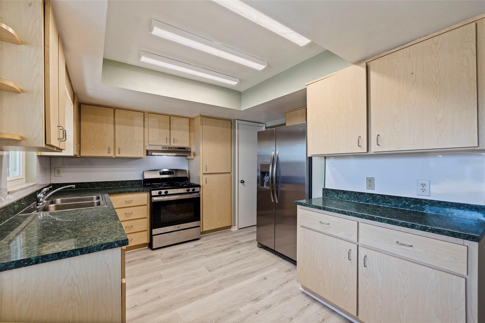 Abundant cabinetry and extended counter space for meal prep and storage. L-shaped layout provides efficient workflow, anchored by a gas range with vent hood and stainless steel refrigerator. Double-basin sink positioned beneath a window allows for natural light, while overhead lighting enhances brightness throughout. Updated vinyl plank flooring completes the clean, functional design.
