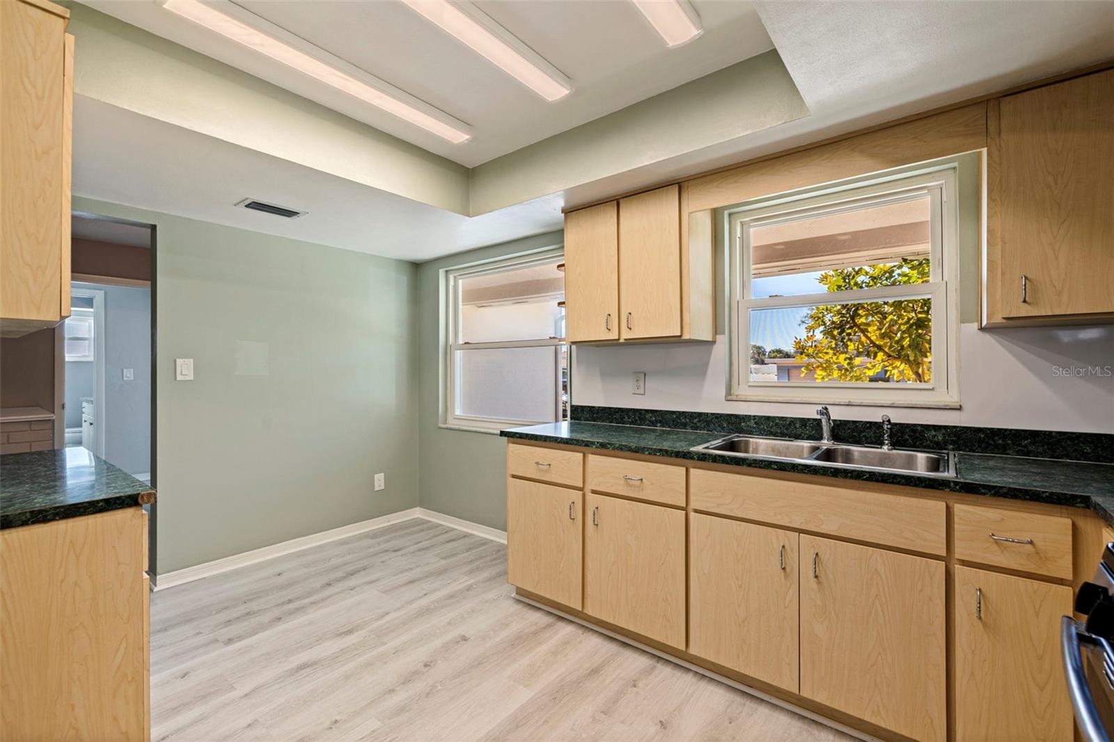 Kitchen with extended cabinetry and generous counter space, highlighted by dual windows that bring in natural light. Double-basin sink positioned beneath the main window offers backyard and neighborhood views. The open corner provides space for a small breakfast table or café-style dining setup. Updated vinyl plank flooring and overhead lighting contribute to a bright, functional layout.