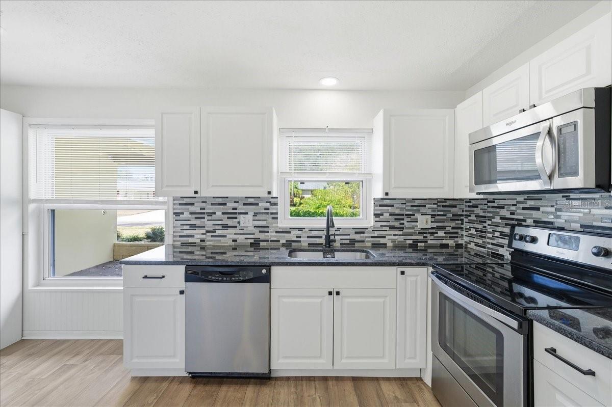 The updated kitchen is bright and welcoming, featuring white cabinetry, two windows, a dinette area, and a prep counter with pendant lighting that opens to the dining area. Black accents and stainless-steel appliances create a crisp, modern look. Ample cabinetry, generous counter space, and direct garage entry combine to create a kitchen designed for both daily living and easy entertaining.