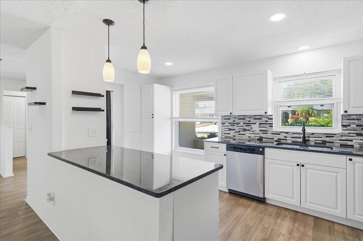 The updated kitchen is bright and welcoming, featuring white cabinetry, two windows, a dinette area, and a prep counter with pendant lighting that opens to the dining area. Black accents and stainless-steel appliances create a crisp, modern look. Ample cabinetry, generous counter space, and direct garage entry combine to create a kitchen designed for both daily living and easy entertaining.