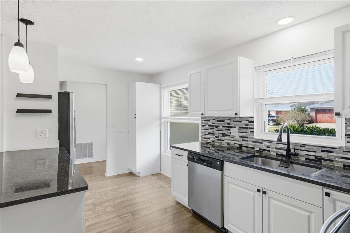 The updated kitchen is bright and welcoming, featuring white cabinetry, two windows, a dinette area, and a prep counter with pendant lighting that opens to the dining area. Black accents and stainless-steel appliances create a crisp, modern look. Ample cabinetry, generous counter space, and direct garage entry combine to create a kitchen designed for both daily living and easy entertaining.