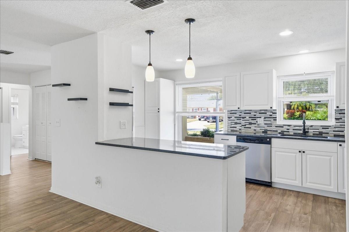 The updated kitchen is bright and welcoming, featuring white cabinetry, two windows, a dinette area, and a prep counter with pendant lighting that opens to the dining area. Black accents and stainless-steel appliances create a crisp, modern look. Ample cabinetry, generous counter space, and direct garage entry combine to create a kitchen designed for both daily living and easy entertaining.