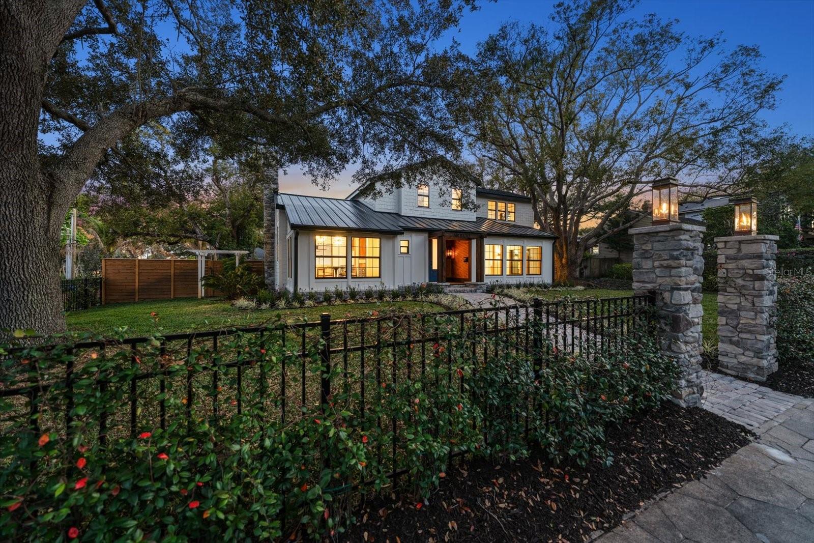 Front of home at twilight with wrought iron fencing and Jasmine. Gas lanterns atop stone pillar walkway entrance