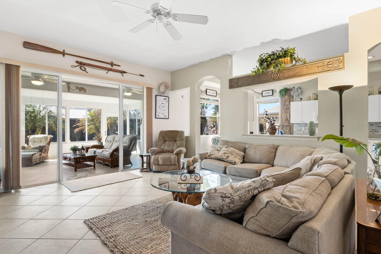 Spacious living area featuring an airy open-concept design, vaulted ceilings, and large-format neutral tile flooring. Oversized sliding glass doors lead directly into the bright sunroom, creating a seamless flow for indoor-outdoor entertaining and flooded with natural Florida sunshine.