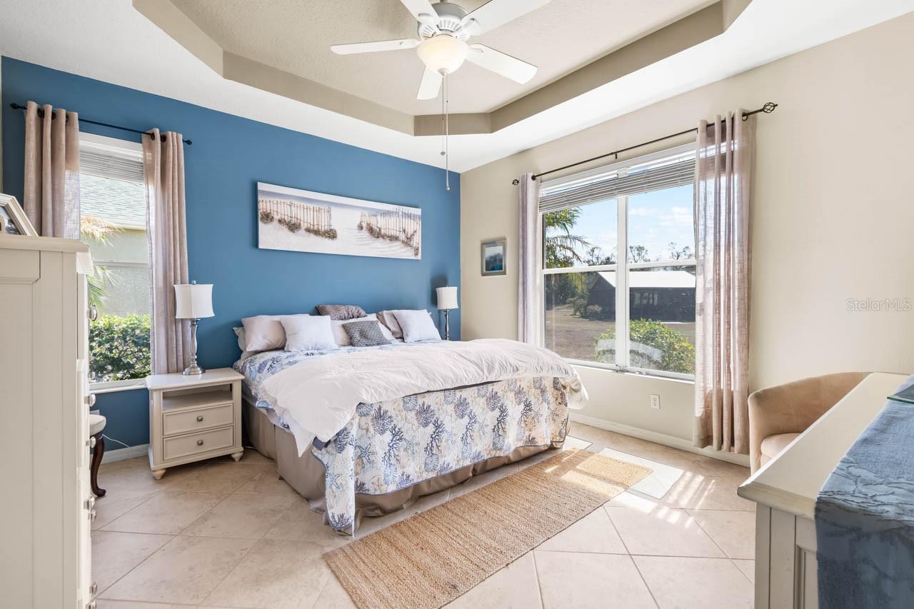 Spacious Primary Bedroom featuring an elegant tray ceiling, stylish coastal blue accent wall, and neutral tile flooring. Large windows provide plenty of natural light and peaceful views of the backyard, while the room easily accommodates king-sized furniture with room to spare.