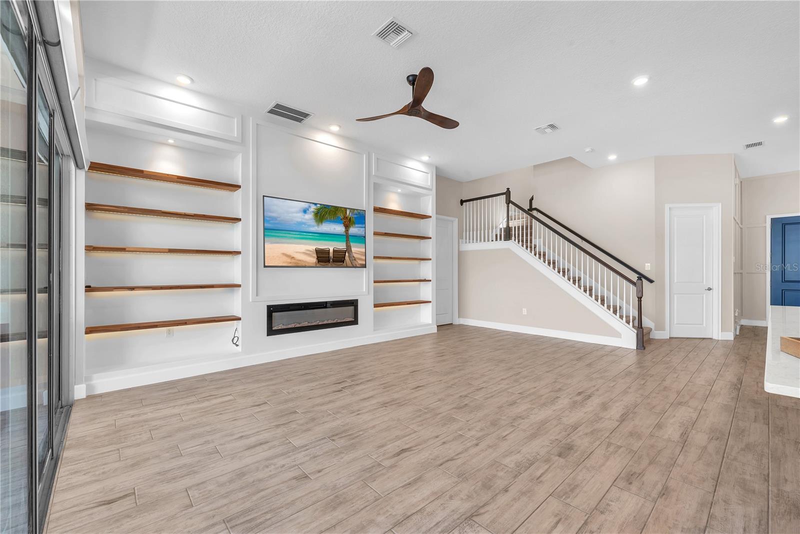 The living room showcases a stunning wall of built-in wood beam shelving with integrated LED lighting, beautifully framing an electric fireplace beneath the large TV (included with the home)