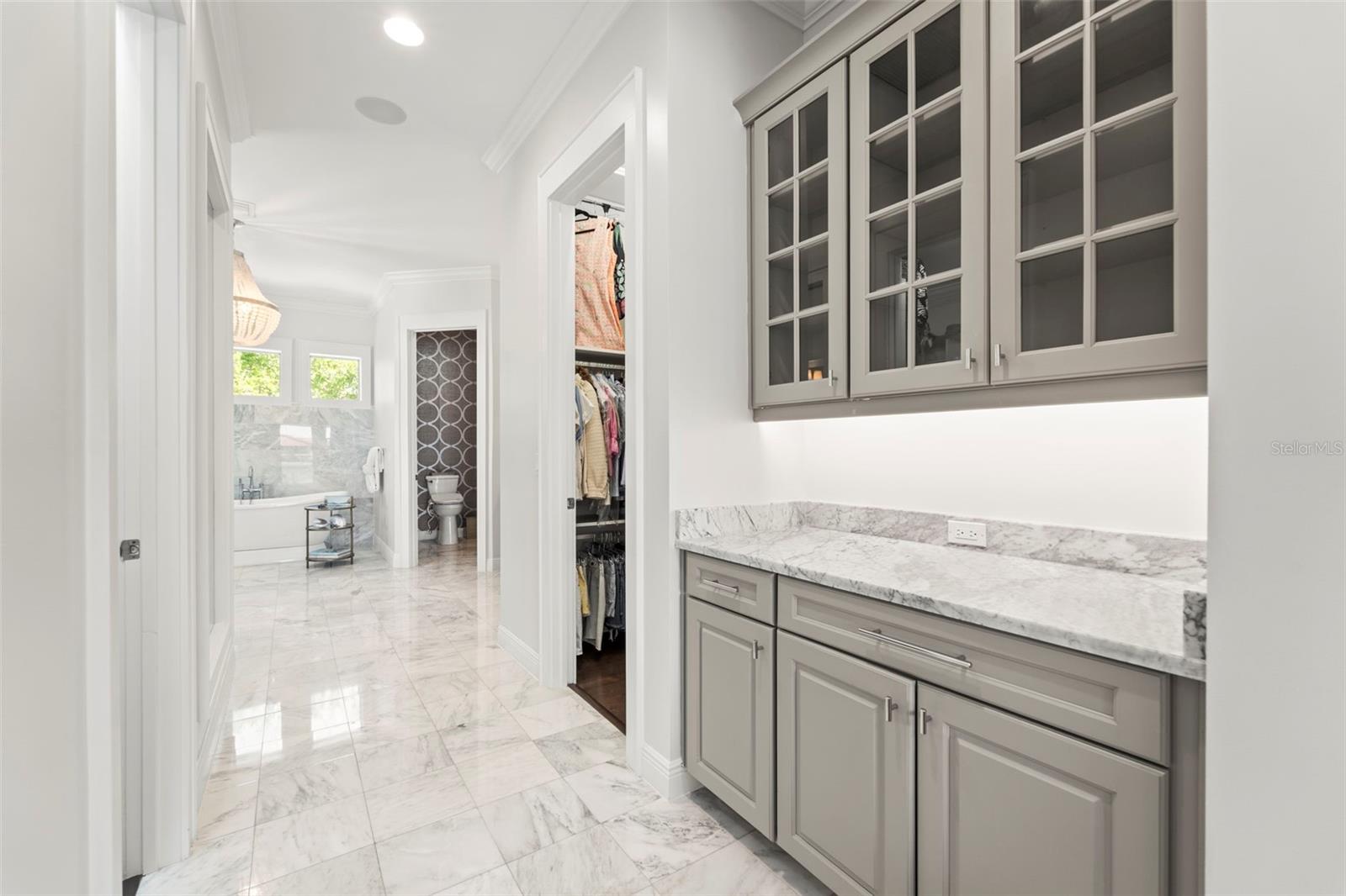 Transition from the bedroom into the spa-inspired primary bathroom, passing a dry bar and two walk-in closets with built-in shelving and storage.
