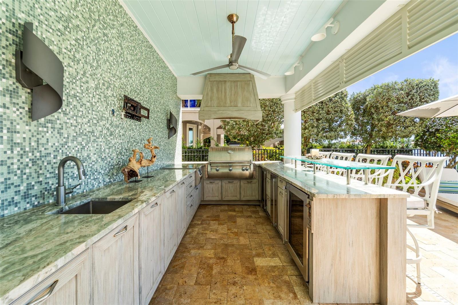 Fully equipped outdoor kitchen featuring built-in grill with exhaust hood, two burner cooktop, sink, ice maker, large built in YETI cooler, dual mini refrigerators, and hidden dual trash bins.