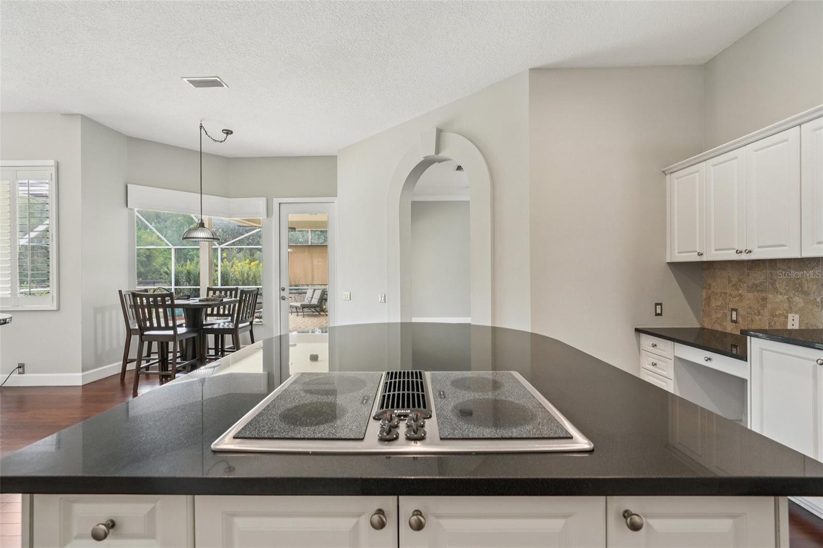 The modern kitchen is thoughtfully designed with a functional layout including an abundance of bright white cabinetry, complementary dark granite countertops a central island with extended overhang and sleek Jenn-Air downdraft cooktop. The premium appliance package also includes stainless steel built-in wall ovens and a French door refrigerator. The eat-in breakfast nook has mitered windows providing expansive views of the pool and conservation, recessed lighting, and a seamless flow into the family room.