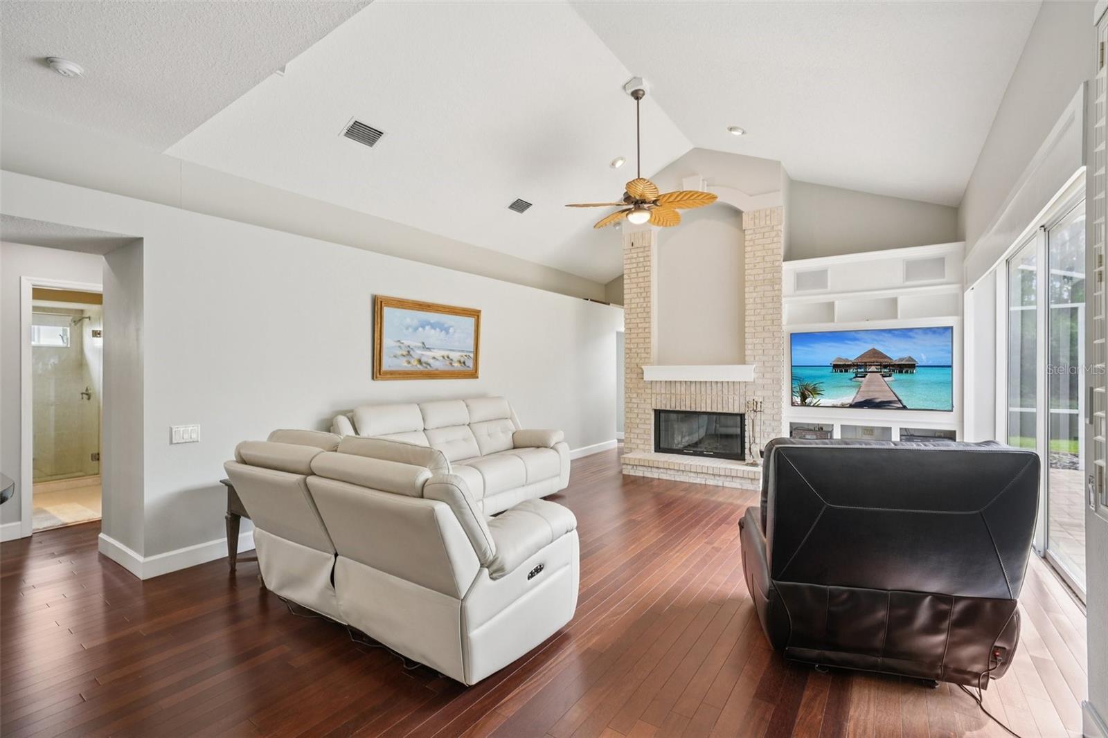 The family room is an ideal gathering space with vaulted ceilings, expansive triple glass sliders opening to the pool area, built-in shelving with media space, and a striking wood-burning fireplace.