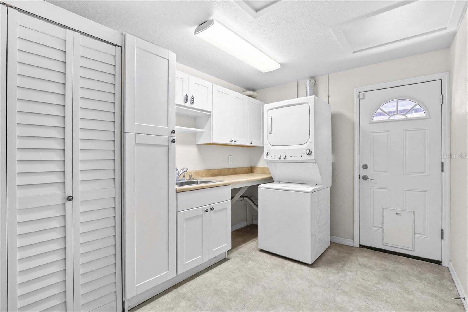 Convenient interior laundry space with white shaker cabinetry providing an abundance of storage, a utility sink, and exterior door to the side yard that could be a fenced dog run and backyard