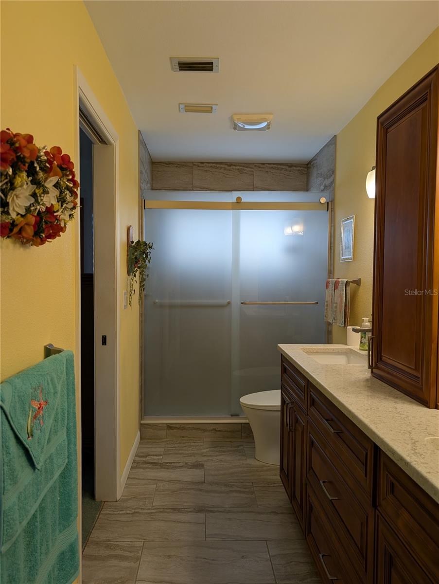 View from interior laundry room into primary bath.  Other notable features include: ceiling vent, walk-in tile shower w/glass doors and comfort height commode.