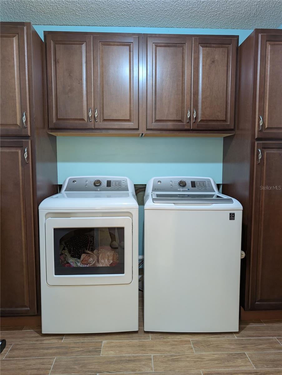 Interior laundry has GE washer and dryer (2018), rich wood cabinetry and wood plank tile flooring.  Dryer vents thru garage (on left) to outside. Hallway is to the right.