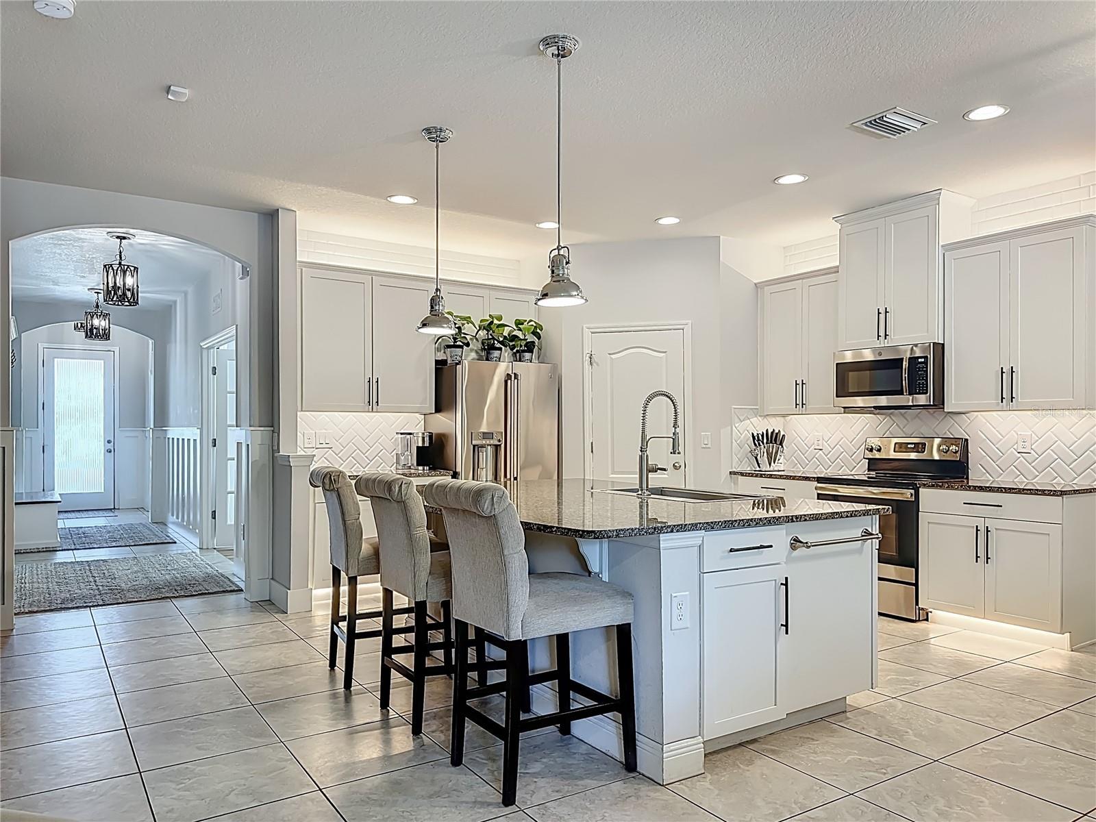 Kitchen boasts white wood cabinetry, granite countertops, a striking herringbone subway tile backsplash, stainless steel appliances, an oversized island with additional cabinetry and a breakfast bar, under-cabinet and over-cabinet LED lighting, a spacious eat-in dining area, and a custom walk-in pantry