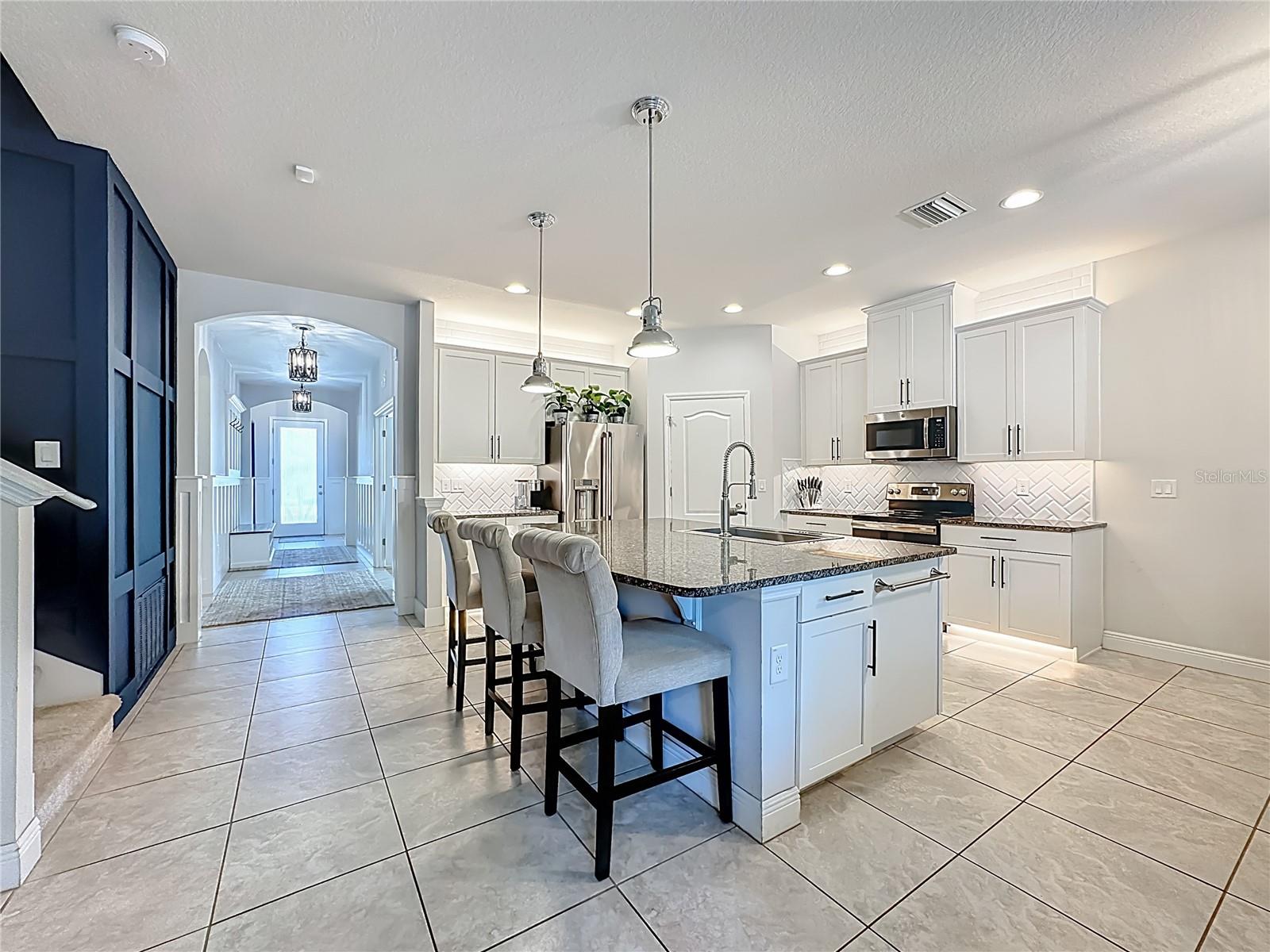 Kitchen boasts white wood cabinetry, granite countertops, a striking herringbone subway tile backsplash, stainless steel appliances, an oversized island with additional cabinetry and a breakfast bar, under-cabinet and over-cabinet LED lighting, a spacious eat-in dining area, and a custom walk-in pantry