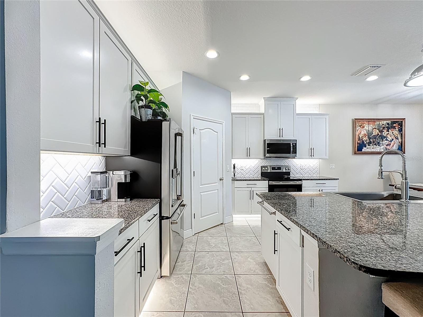 Kitchen boasts white wood cabinetry, granite countertops, a striking herringbone subway tile backsplash, stainless steel appliances, an oversized island with additional cabinetry and a breakfast bar, under-cabinet and over-cabinet LED lighting, a spacious eat-in dining area, and a custom walk-in pantry