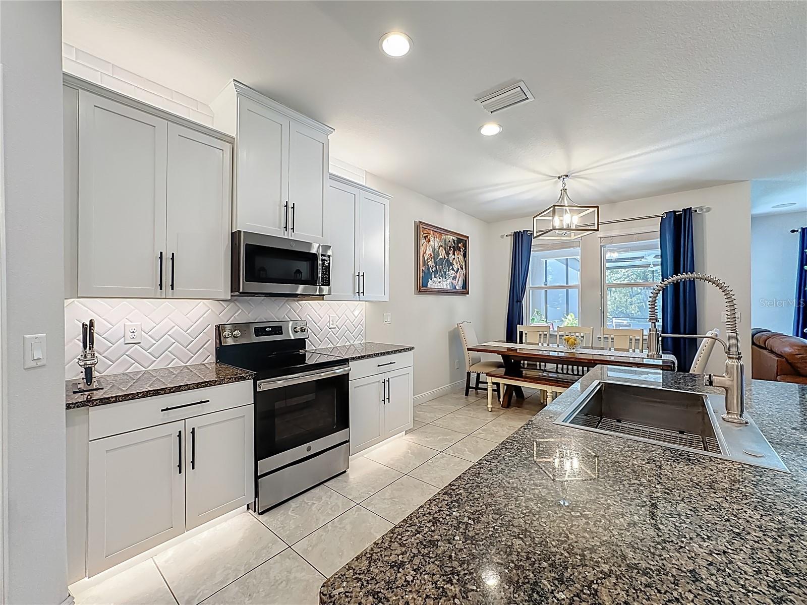 Kitchen boasts white wood cabinetry, granite countertops, a striking herringbone subway tile backsplash, stainless steel appliances, an oversized island with additional cabinetry and a breakfast bar, under-cabinet and over-cabinet LED lighting, a spacious eat-in dining area, and a custom walk-in pantry