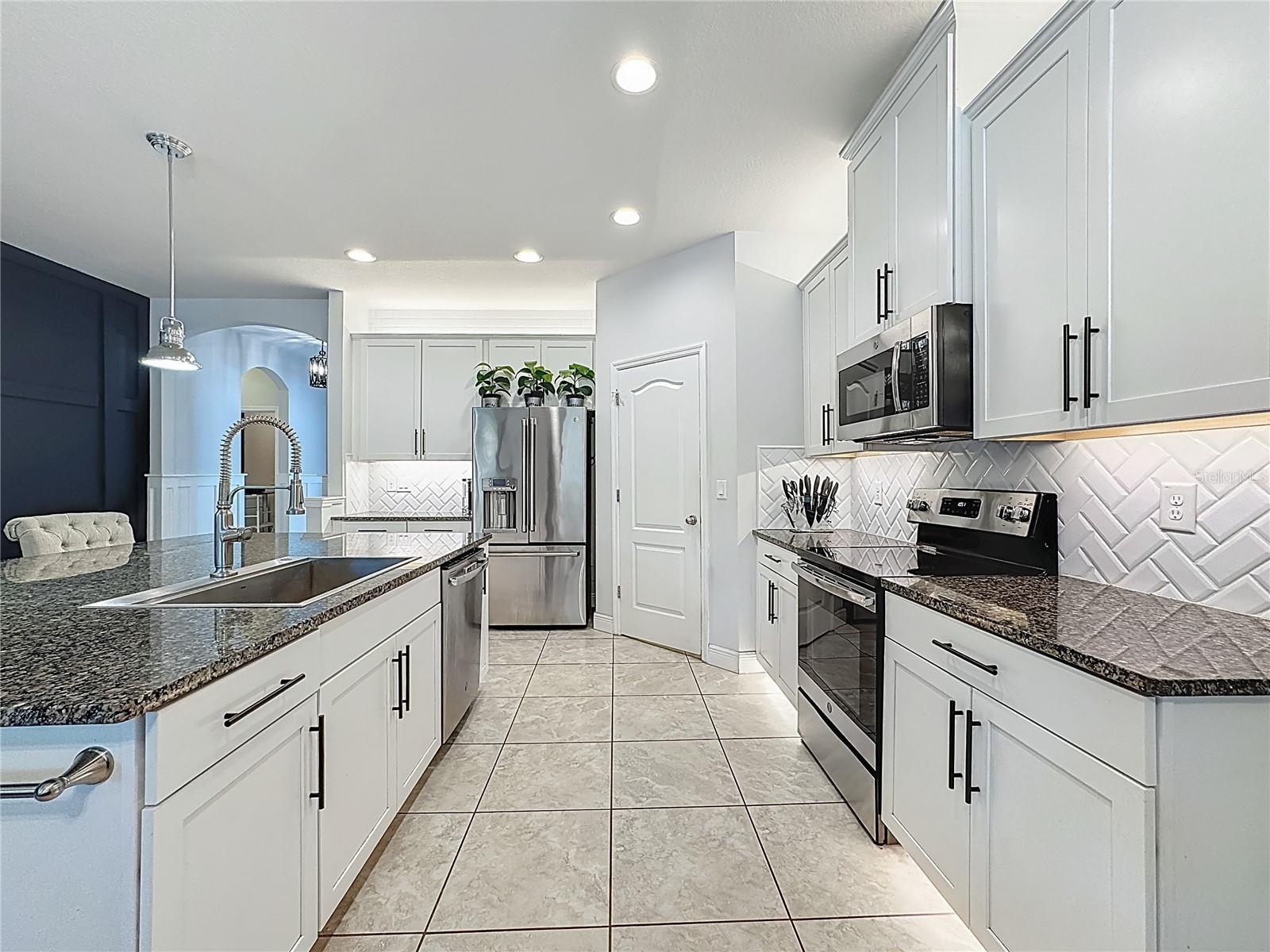 Kitchen boasts white wood cabinetry, granite countertops, a striking herringbone subway tile backsplash, stainless steel appliances, an oversized island with additional cabinetry and a breakfast bar, under-cabinet and over-cabinet LED lighting, a spacious eat-in dining area, and a custom walk-in pantry